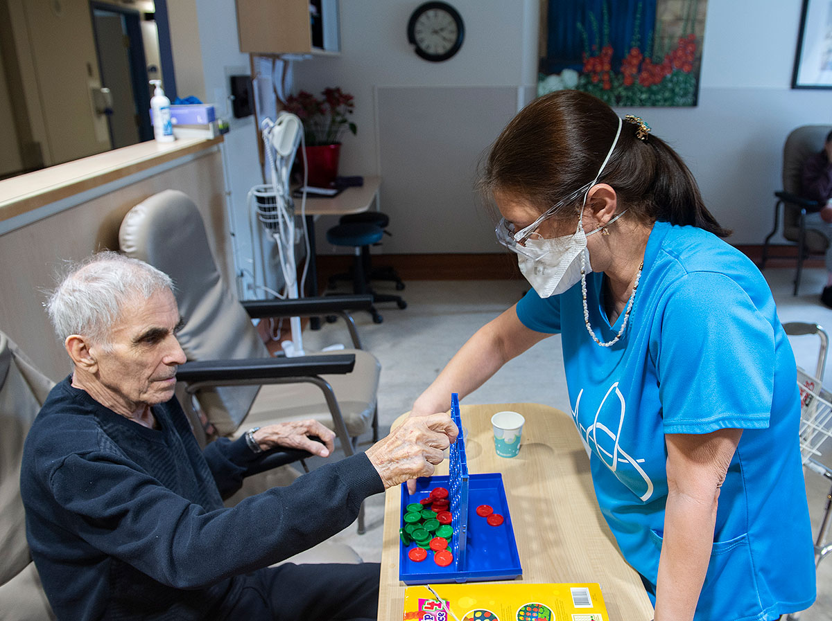A woman wearing a blue nurses uniform plays Connect Four with an elderly man.