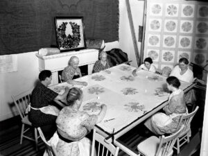 A black and white photo of a group of women sitting around a table sewing a quilt.