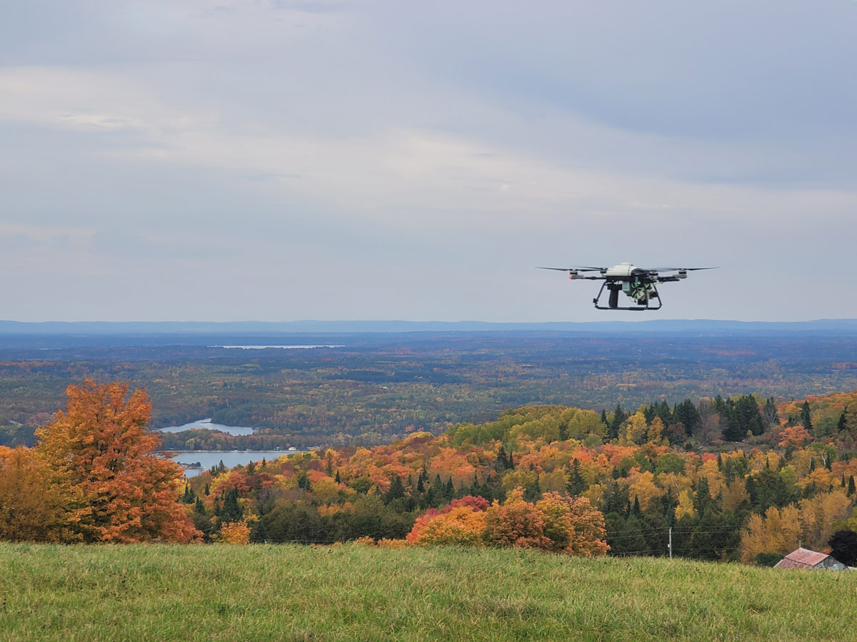 A remotely piloted long-range survey drone flies above an expansive area of deciduous and coniferous trees in autumn and some small bodies of water with a blue sky