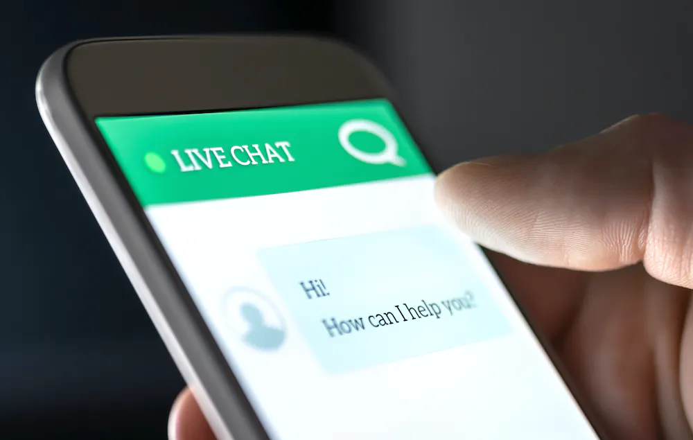 A hand holding a phone screen showing a live chat with the text HI HOW CAN I HELP YOU?