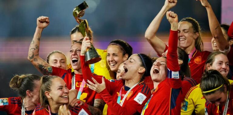 A women's World Cup winning team in red jersey's hold up their trophy and cheer