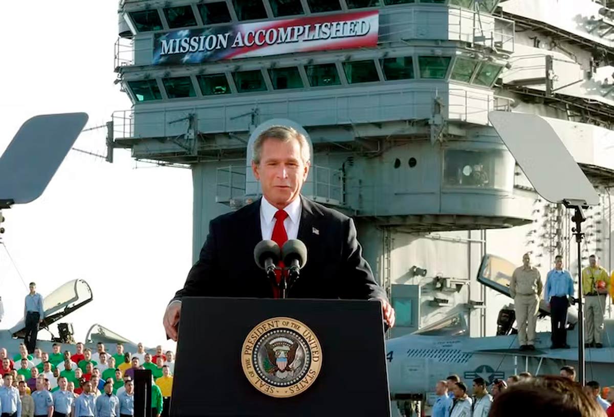 A grey-haired man stands a podium with the U.S. presidential insignia. Behind him a sign reads Mission Accomplished.