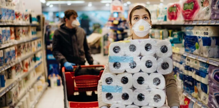 A person wearing a mask and holding tissue paper rolls in the foreground and person wearing a mask while holding a shopping cart on the background