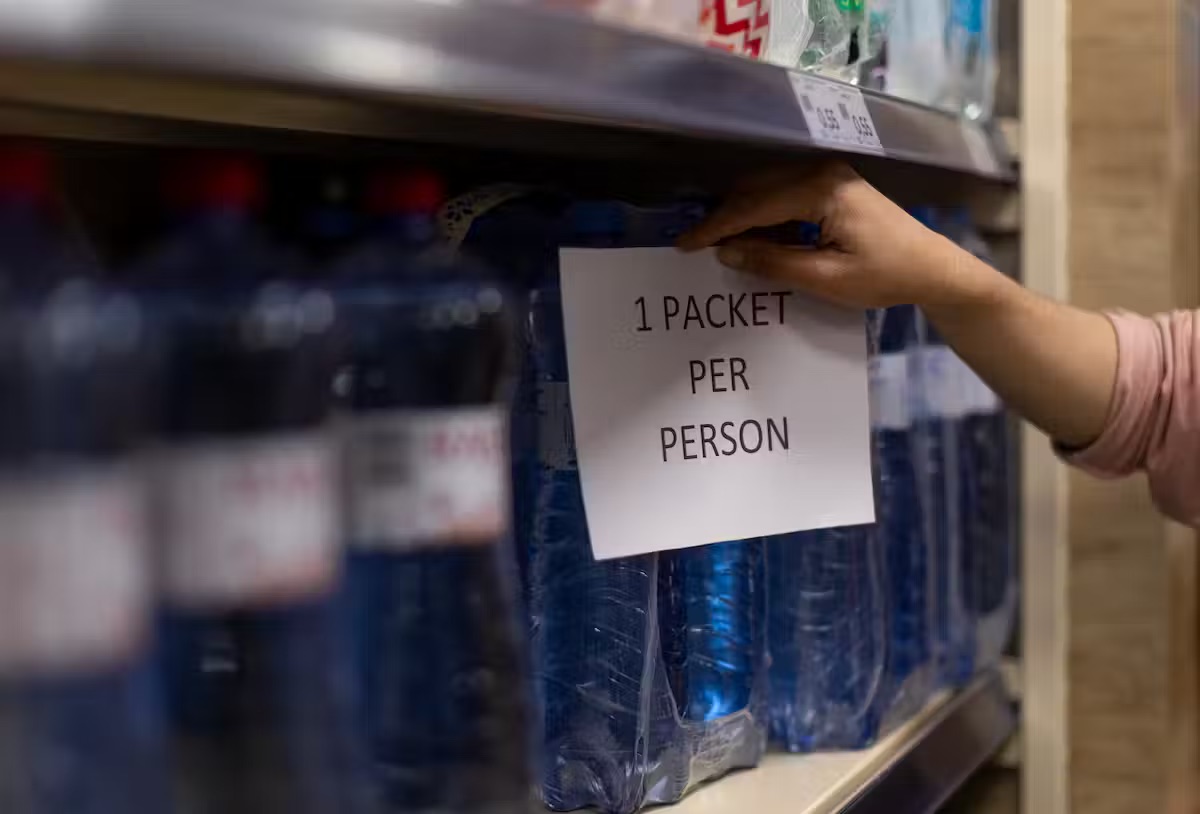 A sign that says '1 packet per person' taped to a shelf of water bottles"