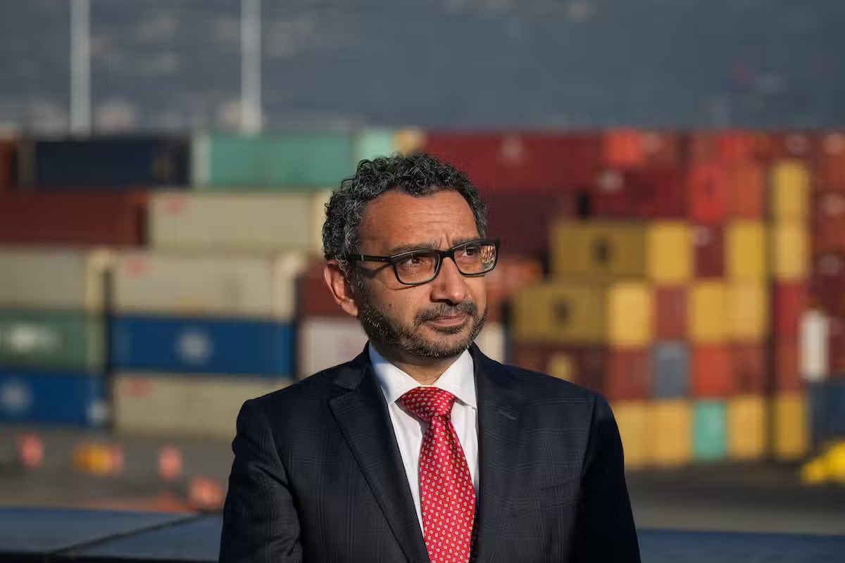 A South Asian man in a black suit and red tie stands in front of a port full of large supply crates.