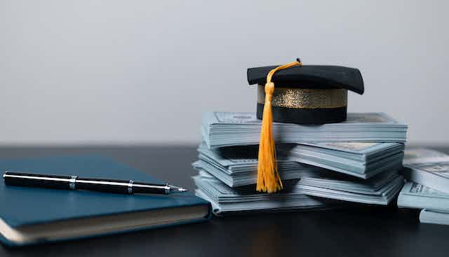 A graduation cap sits on a stack of money next to a book and pen.