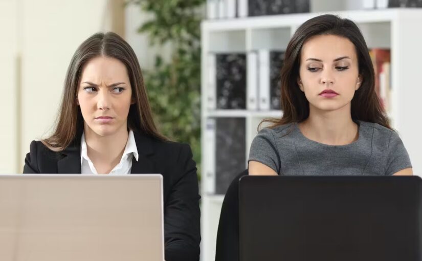 Two women give each other jealous looks while working on laptops.