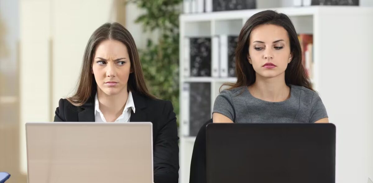 Two women give each other jealous looks while working on laptops.