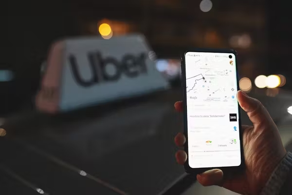 Uber app displayed on smartphone held in a hand in front of a taxi with an Uber sign on top of a car.