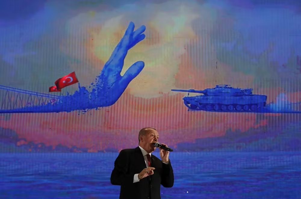 A man in a dark suit speaking on in front of a large image of a raised hand with the Turkish flag facing a military tank.
