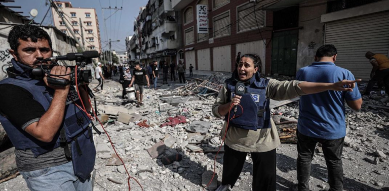 A journalist reports amid the destruction and rubble in Gaza while a cameraman records.