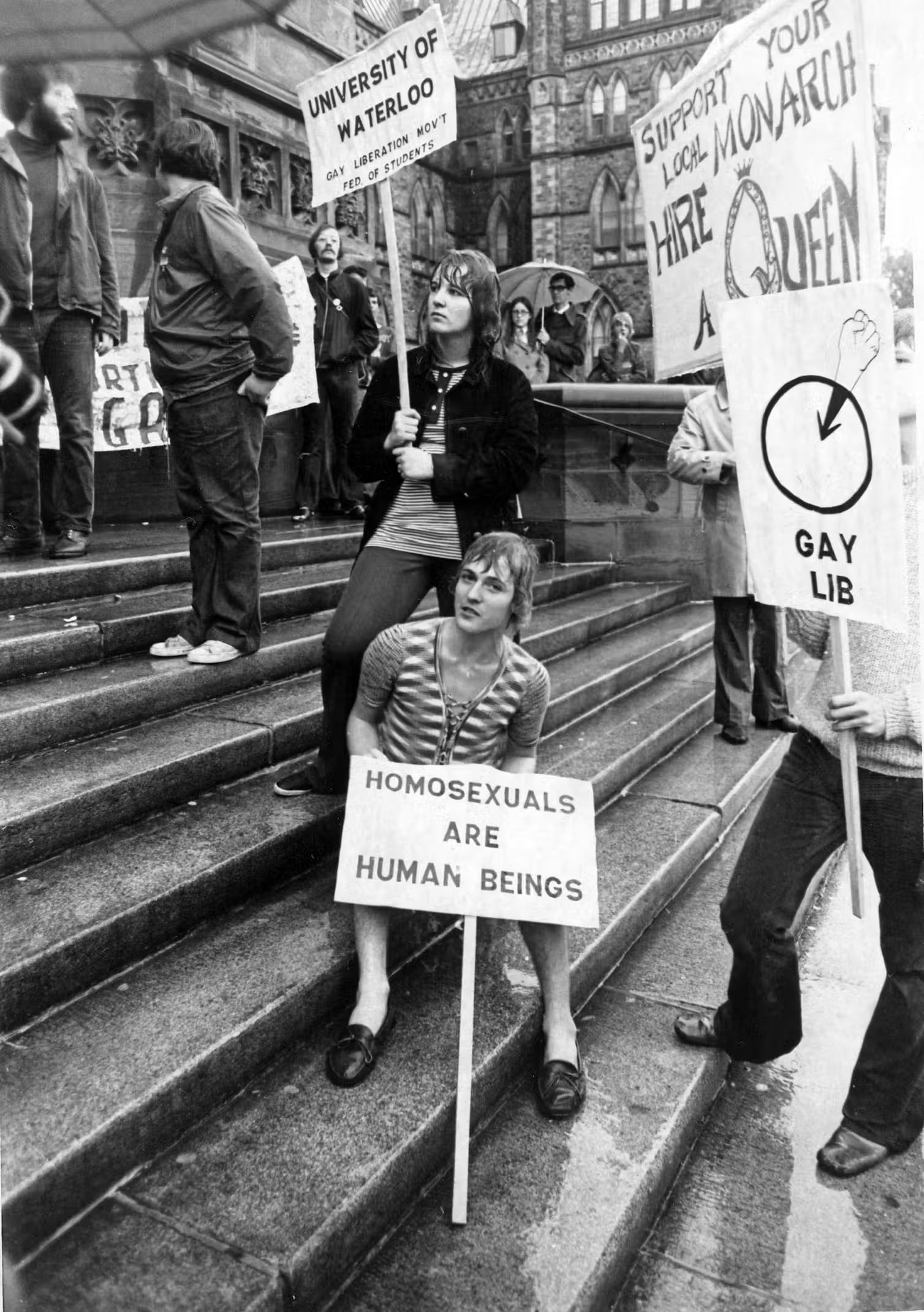 A black and white photo of people protesting on building steps. One placard reads: homosexuals are human beings.