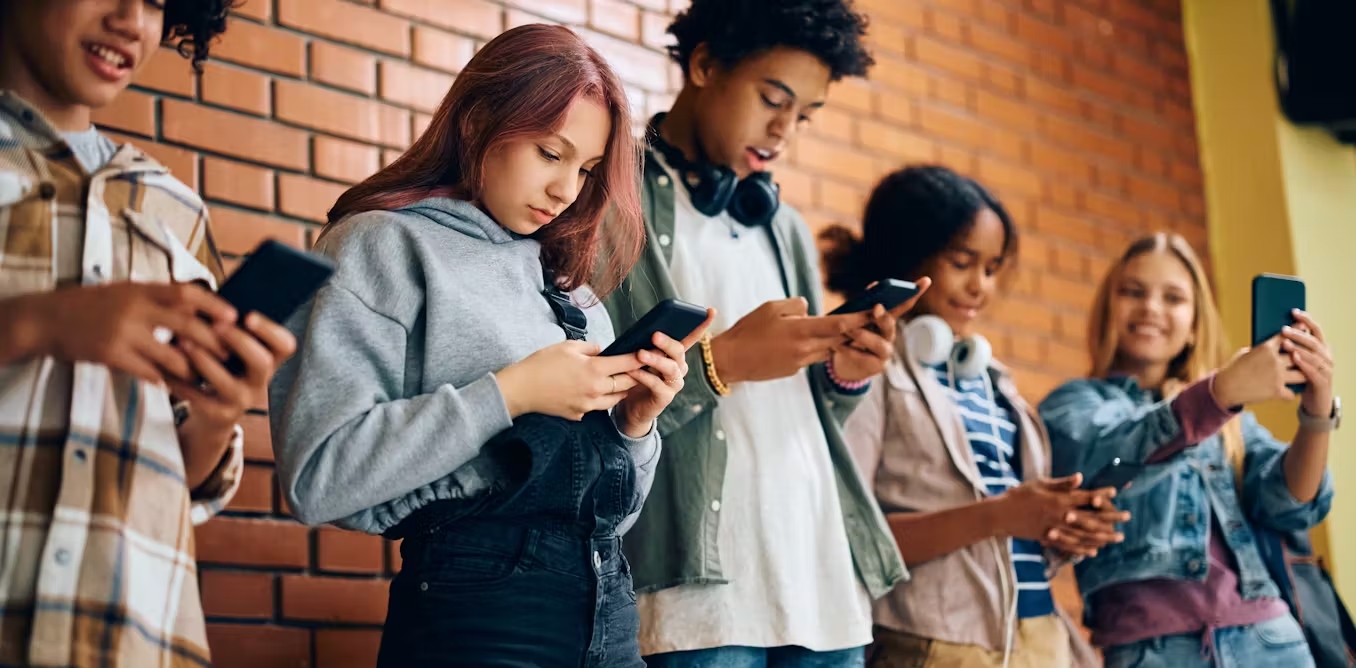 A group of teenagers look at their phones while standing in front of a brick wall.