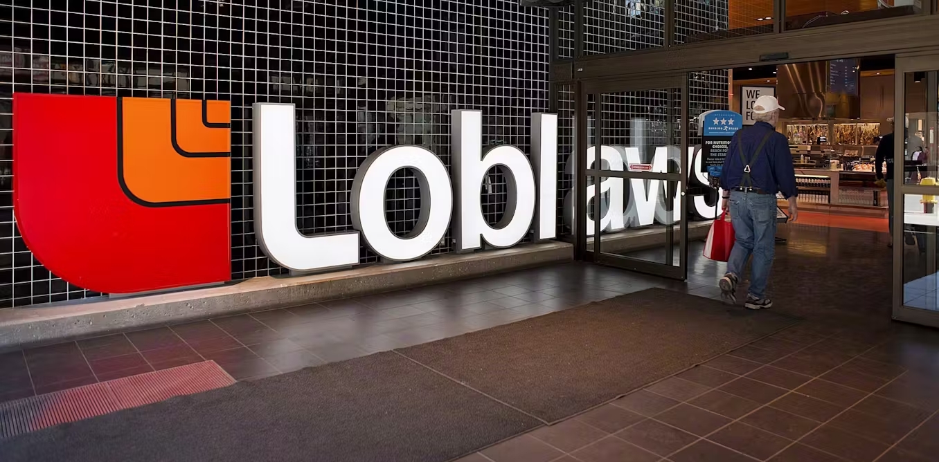 A man carries a bag of groceries while walking in front of a Loblaws sign.