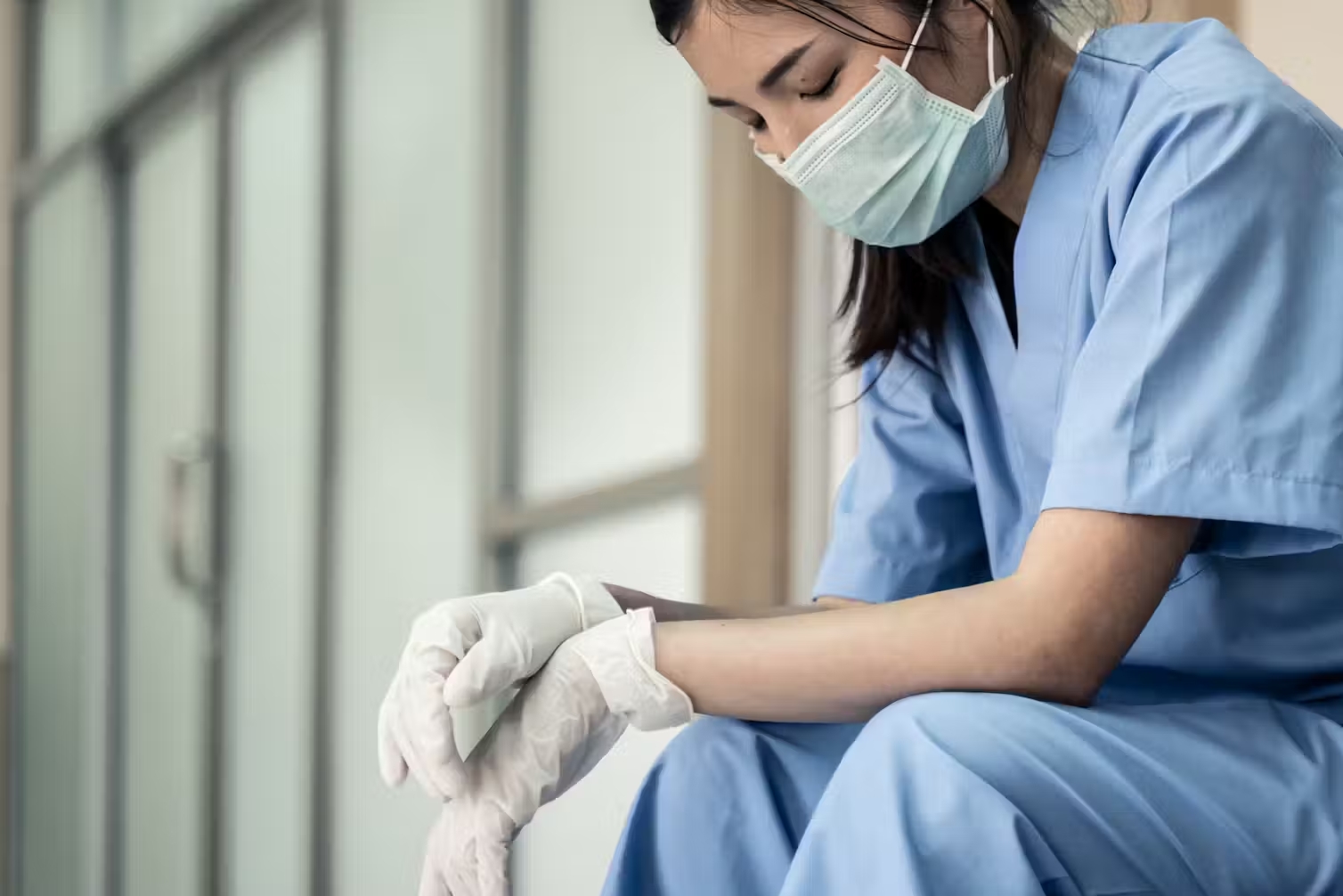A health-care worker in blue scrubs and a face mask looking tired.
