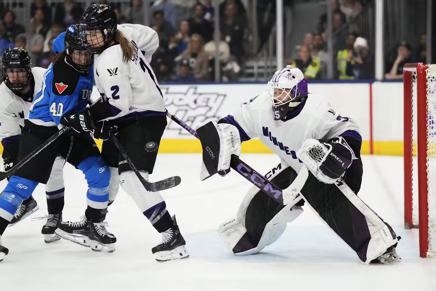 A hockey goalie crouches with their stick out as two other hockey players fight over a puck beside them.