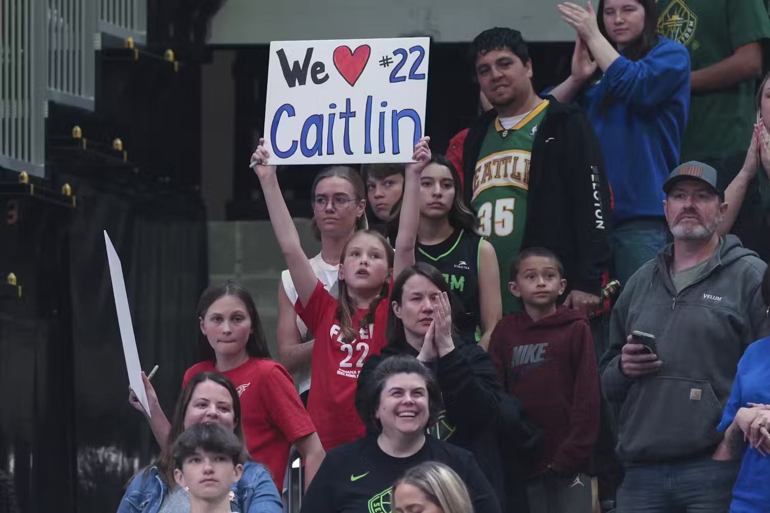 A young girl sitting in the stands of a sport stadium holds up a sign that says 'We love Caitlin #22'