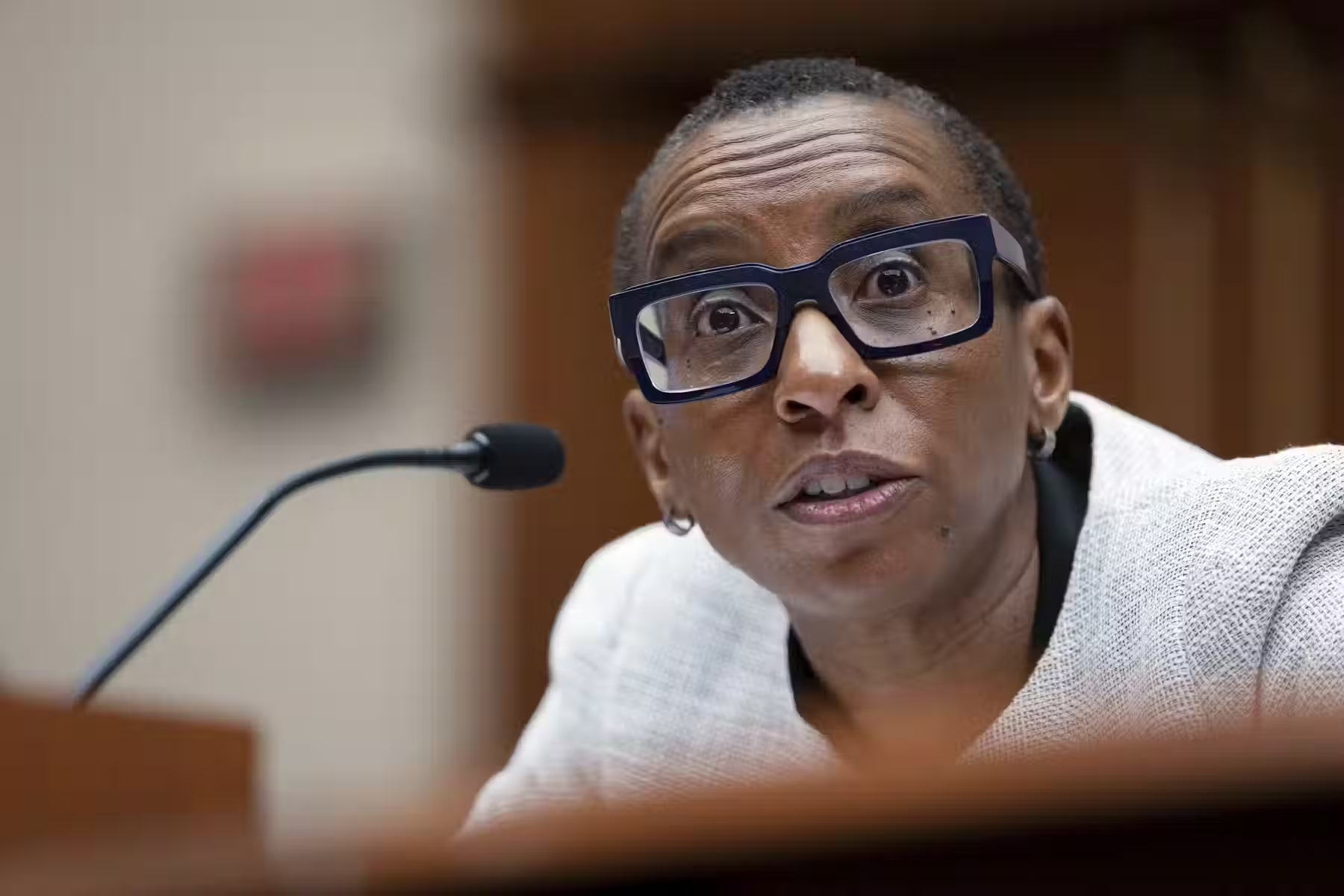 A Black woman with short hair and wearing glasses speaks into a microphone.