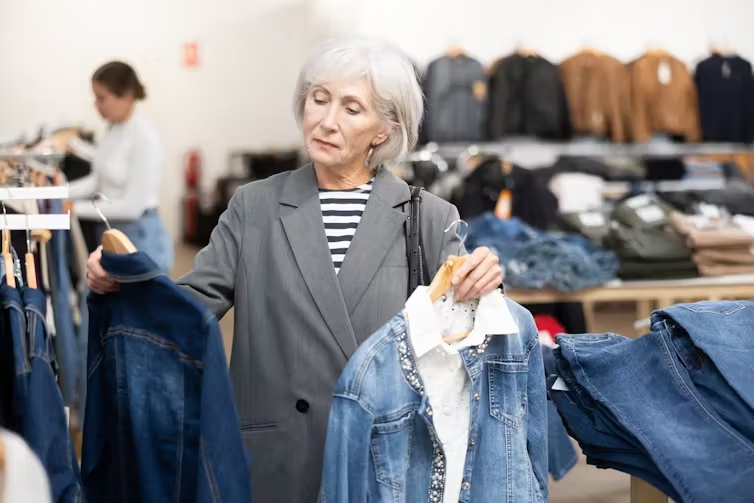 An older woman holds two hangers with denim jackets on them, one in each hand, while standing before a rack of clothes in a store.