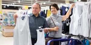An older man holds up two shirts on hangers, one in each hand, while a woman reaches for a shirt on a clothing rack in a store.